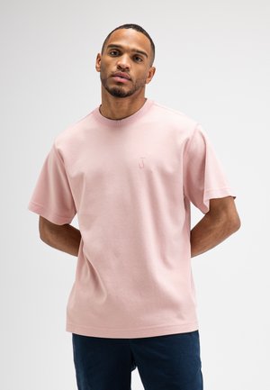 Man met kort haar en baard die een lichtroze t-shirt met korte mouwen en donkere broek draagt, staand met handen achter de rug.