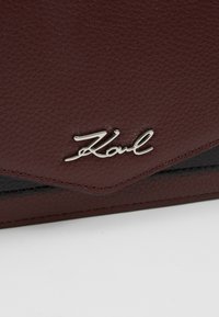 Burgundy struktureret lædertaske med sorte detaljer, prydet med et sølvlogo. Sammenfoldt design med en glat finish og skarpe kanter.