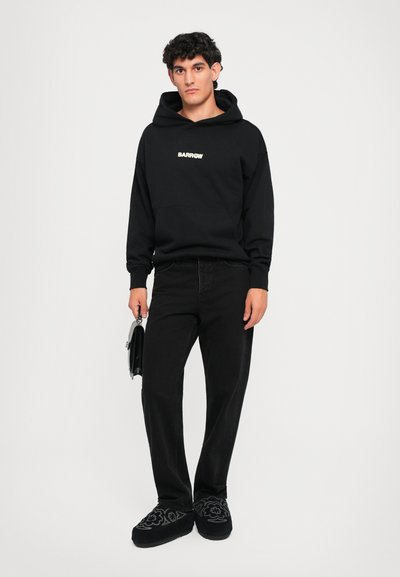 Sudadera negra con bolsillo frontal y logo blanco, combinada con jeans negros de pierna recta y zapatos negros de lona bordados. Sosteniendo un bolso pequeño.