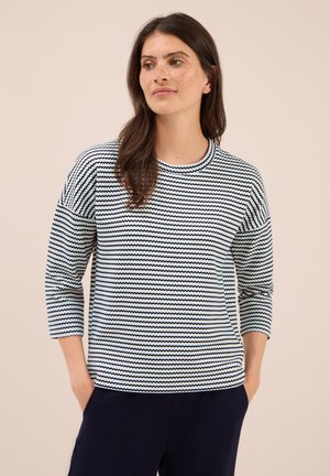 Donna con lunghi capelli castani che indossa un maglione a righe ondulate bianche e nere e pantaloni scuri, con le mani in tasca, che guarda leggermente di lato.
