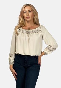 Beżowa bluzka z koronkowym wykończeniem dekoltu i rękawów, z obszernymi rękawami i luźnym krojem, zestawiona z ciemnymi dżinsami.