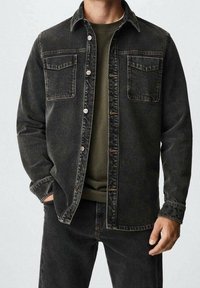Chaqueta de mezclilla negra con dos bolsillos en el pecho, botones en la parte frontal y un corte relajado, llevada sobre una camisa verde oscura, con detalles de costura visibles.