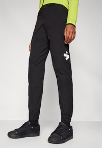Svart, lette bukser med tett passform, glidelåslommer og en hvit logo på venstre ben. Kombinert med svarte teksturerte joggesko.