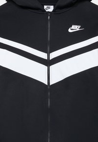 Juodas zip-up hoodie su baltomis chevron juostelėmis ir Nike logotipu ant krūtinės. Medžiaga yra lygi ir minkšta.