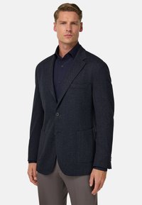 Granatowy blazer w klasyczny wzór jodełki, wykonany z teksturowanej tkaniny, z pojedynczym zapięciem na guzik, klapami w kształcie notcha oraz dwoma przednimi kieszeniami. Można go zestawić z ciemną koszulą.
