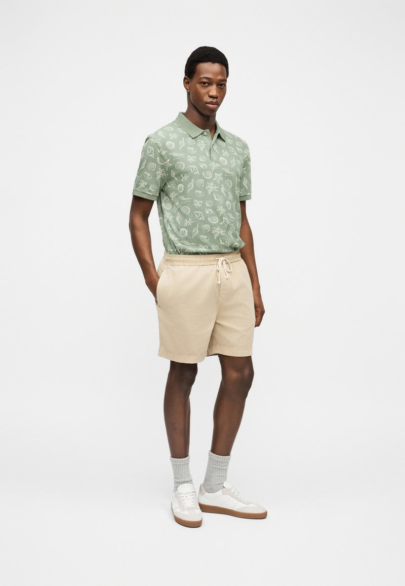 Uomo in piedi con le mani nelle tasche, indossa una polo verde a motivo, pantaloncini beige, calzini grigi e scarpe da ginnastica bianche su sfondo semplice.