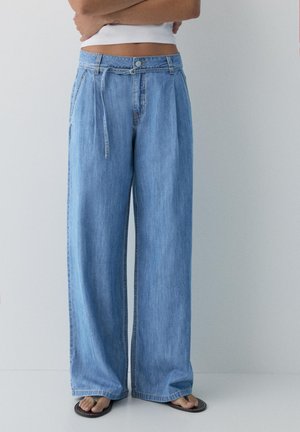 Személy világoskék, magas derekú, bő szárú farmernadrágot visel textil övvel, fehér rövid felsővel és fekete flip-flop papuccsal.