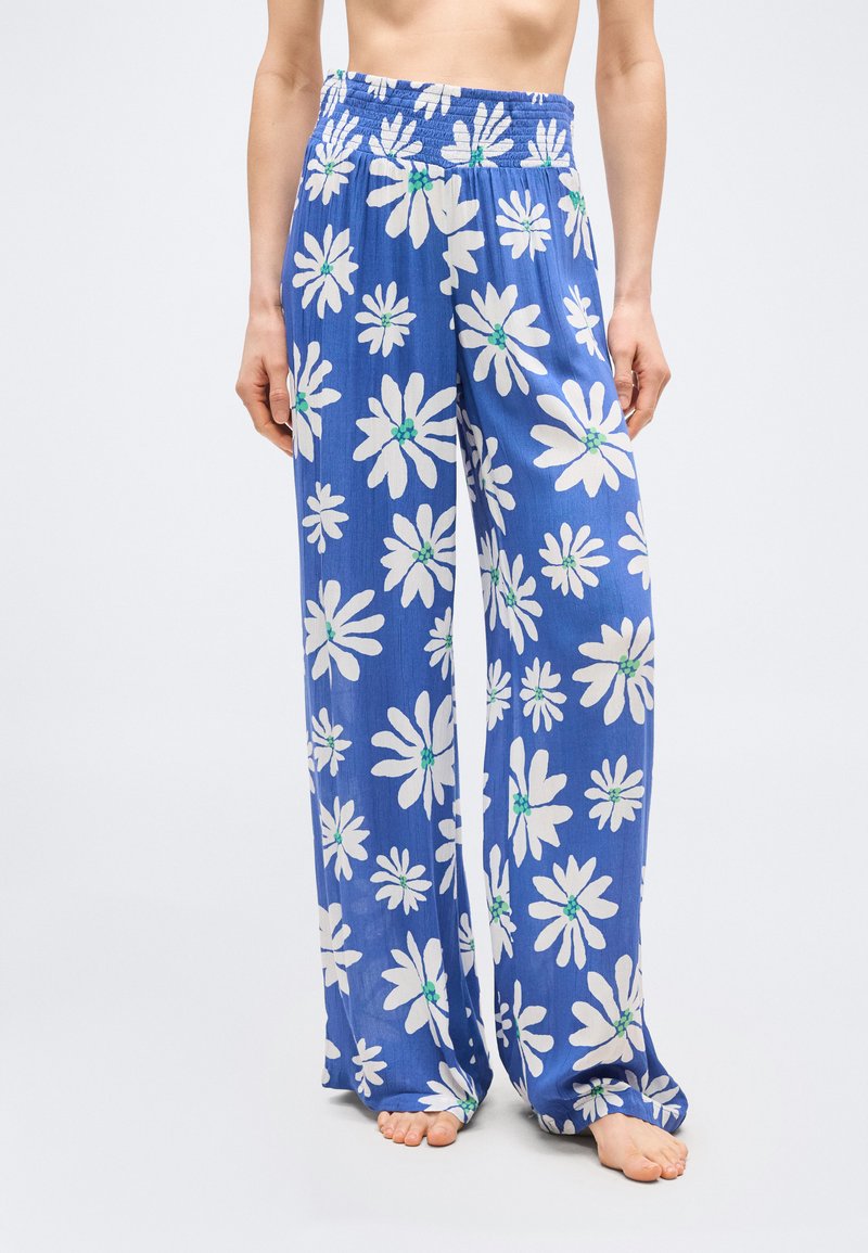 Γυναίκα που φορά χαλαρά μπλε παντελόνια με μεγάλα λευκά σχέδια μαργαρίτας, όρθια ξυπόλυτη μπροστά σε απλό λευκό φόντο.