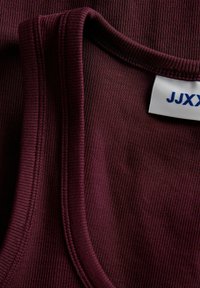 Gros plan d’un tissu côtelé bordeaux foncé plié montrant la couture de l’encolure et une étiquette de vêtement blanche partielle avec un texte bleu.