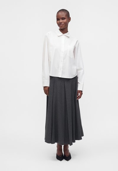 Personne portant une chemise blanche boutonnée et une longue jupe plissée gris foncé, debout devant un fond clair uni.