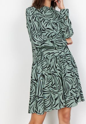 Vrouw draagt een loszittende, langarmige groene jurk met zwarte abstracte zebra-achtige strepen, poseert met één hand bij haar gezicht.