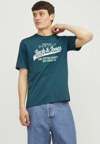Teal bomulds T-shirt med hvidt trykt logo og tekst. Korte ærmer, rund hals, afslappet pasform, parret med lyseblå denimjeans.
