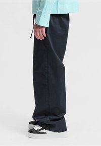 Personne portant un pantalon noir à jambes larges, des baskets noires et blanches, et une chemise à manches longues rayée bleue, debout de profil sur un fond blanc.