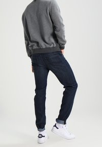 Dunkelblaue Jeans mit geradem Schnitt, kombiniert mit einem grauen Sweatshirt und weißen Turnschuhen. Sichtbare Nähte an den Jeans und umgeschlagene Säume.