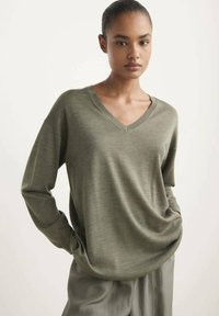Pull à col V vert olive avec des manches longues, fabriqué en tissu doux, arborant une coupe décontractée et une texture subtile. Assorti avec un pantalon de couleur claire.