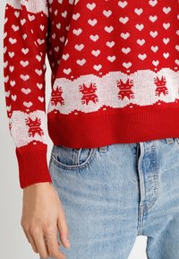 Pull en tricot rouge avec des motifs de cœurs blancs, une bande décorative blanche avec des accents rouges, des poignets côtelés et une coupe décontractée.