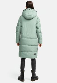 Long, mint green padded coat with a large hood, featuring black lettering on the collar, worn over dark jeans and black lace-up boots.