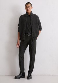 Cardigan en tricot gris foncé avec un col haut, fermeture à boutons, associé à une chemise noire et un pantalon noir ajusté ; chaussures noires brillantes.