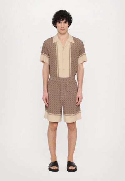 Ensemble chemise à manches courtes et short en beige et marron avec des motifs géométriques, taille élastique et complété par des sandales à glissière noires.