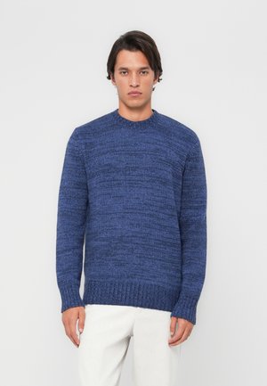 Navy blue knitted sweater with a textured pattern, round neckline, and ribbed cuffs. Worn with light-colored trousers.