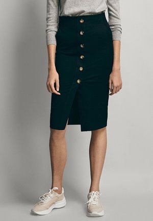 Femme portant une jupe crayon noire à boutons sur le devant, un haut gris à manches longues et des baskets beiges, debout devant un fond gris uni.