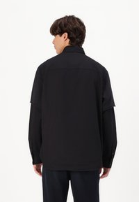 Cămașă neagră cu nasturi, cu o croială lejeră, cu mânecă lungă și detaliu la spate. Textură netedă, design simplu și fără modele vizibile.