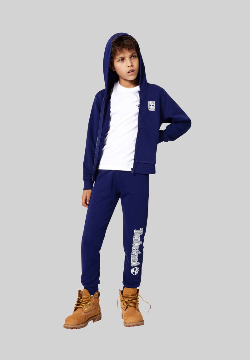 Marineblauwe sweatshirt en broek set van zacht materiaal, voorzien van een rits, capuchon en Timberland-logo. Draagt het met tankleurige laarzen, wat de look accentueert.