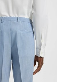 Light blue tailored trousers with pleats, featuring two back pockets and a smooth fabric texture, paired with a white long-sleeve shirt.