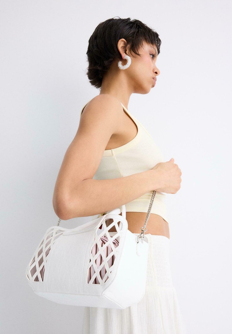 Femme aux cheveux courts et foncés portant un crop top crème et une jupe, tenant un sac à main blanc avec un motif en treillis et une bandoulière en chaîne argentée sur l'épaule.
