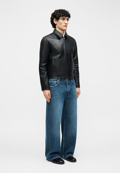 Νεαρός άνδρας με σγουρά μαλλιά, που φοράει μαύρο δερμάτινο μπουφάν, τζιν με φαρδιά παντελόνια και μαύρα παπούτσια, στέκεται μπροστά σε ένα απλό φωτεινό φόντο.