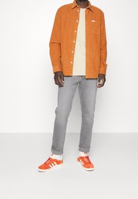 Camisa de pana naranja con botones sobre una camiseta beige, combinada con jeans ajustados grises y zapatillas de ante naranja con rayas blancas.