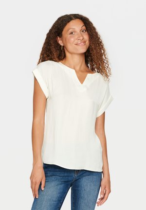 Vit kortärmad blus med V-hals, med uppvikta ärmar och ett mjukt, texturerat tyg, matchad med blå tajta jeans.