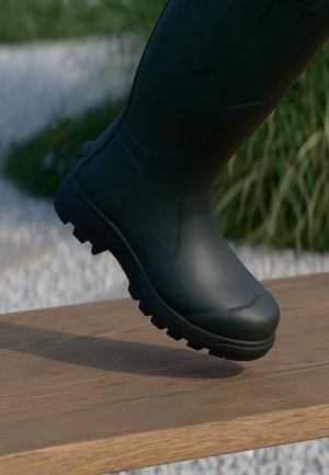 Botte en caoutchouc noire avec une surface texturée, semelle robuste et crans surélevés, montrée en train de poser sur une planche en bois. L'arrière-plan présente de la verdure.