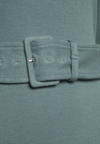 Lähikuva tekstuuriroheisesta kankaisesta vyöstä, jossa on kangaspäällysteinen suorakulmainen solki ja ommellut vyöreiät.