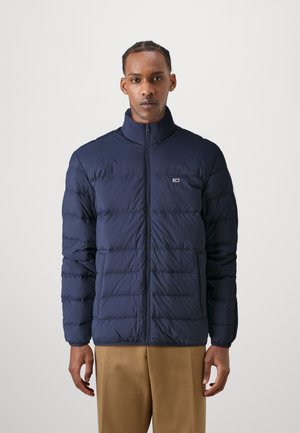 Chaqueta de plumón azul marino con cuello alto, con segmentos acolchados, cremallera frontal y un pequeño logo en el pecho. Textura suave.