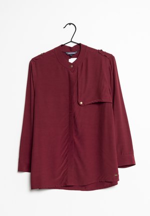 Blusa bordeaux a maniche lunghe con scollatura rotonda, chiusura frontale con bottoni e una tasca con sottotassa fissata da un bottone color oro. Texture liscia.