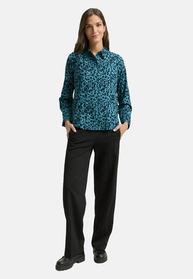 Blaues Hemd mit Leopardenmuster und Knöpfen, lange Ärmel, schwarze Hose und klobige schwarze Schuhe. Glatter Stoff und maßgeschneiderte Passform.