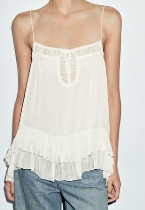 Kvinde iført en hvid chiffonbluse uden ærmer med blonde detaljer og flæsekant, kombineret med blå denimjeans.