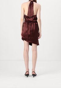 Femme debout, dos à la caméra, portant une robe en satin bordeaux sans manches à col halter et des talons noirs sur fond blanc uni.
