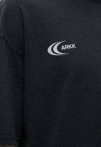 Μαύρο ύφασμα με λευκό λογότυπο ARKK που απεικονίζει δύο καμπύλες γραμμές κοντά στην περιοχή του ώμου.