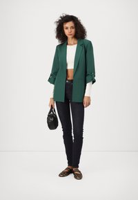 Grün langer Blazer mit weiten Ärmeln, getragen über einem weißen Cropped-Top, kombiniert mit dunklen Skinny-Jeans und schwarzen gemusterten Ballerinas. Schwarze Handtasche inklusive.