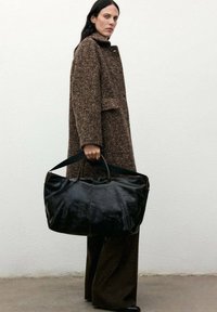 Gran bolso tote de cuero negro grande con dos asas, que presenta una textura suave y costuras visibles. Se lleva con un abrigo texturizado marrón y pantalones.