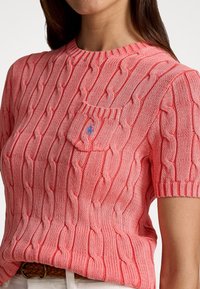 Pull court en tricot torsadé à manches courtes de couleur rose clair, avec une poche ornée d'un emblème bleu. Motif texturé sur l'ensemble du tissu.