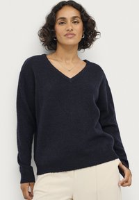 Marineblauer V-Ausschnitt-Pullover aus weichem, strukturiertem Stoff mit langen Ärmeln und lockerer Passform, kombiniert mit hellen Hosen.