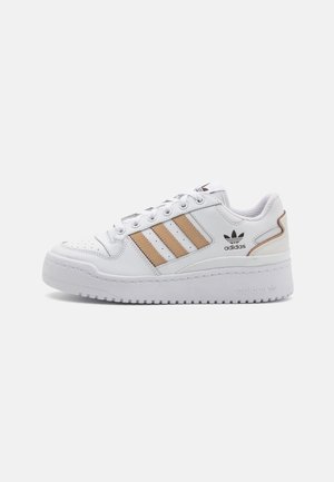 Hvid Adidas-sneaker med beige striber, sort Adidas-logo, perforeret tå, tyk sål og snørelukning mod en ensfarvet baggrund.