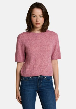 Frau mit kurzen braunen Haaren, die einen pinkfarbenen Kurzarmpullover und blaue Jeans trägt, steht vor einem schlichten hellen Hintergrund.