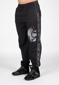 Zwarte, gestructureerde sweatpants met een grafisch ontwerp van een gorilla aan de ene zijde, voorzien van een witte verticale streep en zakdetails. Zwarte sportieve schoenen.