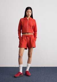 Veste courte zippée rouge et shorts assortis en tissu texturé, complétés par des chaussures rouges et des chaussettes blanches. Design et coupe simples.