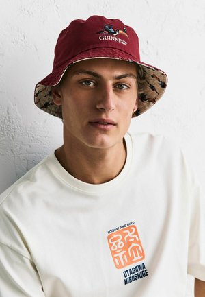 Junger Mann mit rotem Guinness-Bucket-Hut und weißem T-Shirt mit orange-blauem grafischem Text auf der Brust, vor einer strukturierten weißen Wand.