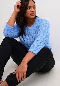 Helles blaues Strickpullover mit einem strukturierten offenen Muster und gerippten Bündchen, kombiniert mit schwarzen Hosen. Modell sitzt vor einem neutralen Hintergrund.